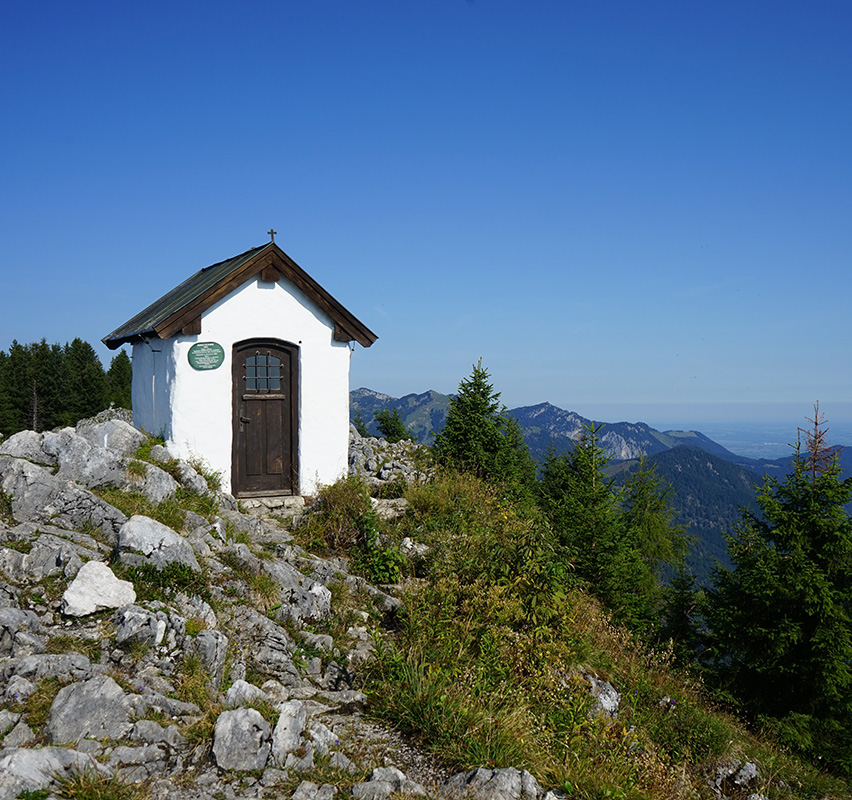 Weiße Kapelle auf einem felsigen Berg mit Kreuz im Vordergrund.