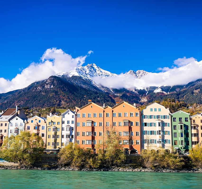 Bunte Gebäudefassade in Innsbruck mit Inn-Fluss und schneebedeckten Bergen.