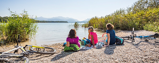 Radfahren_Chiemsee Frauengruppe sitzt auf Kies am See und schaut aufs Wasser.
