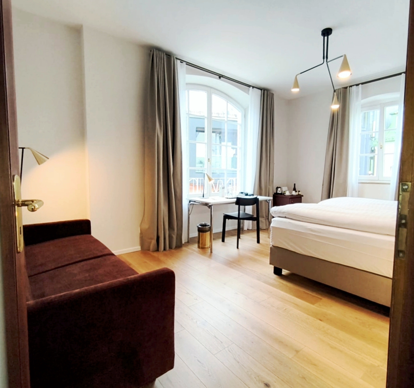 Helles Komfortzimmer im Hotel Das Lindner in Bad Aibling mit großem Doppelbett, Sitzsofa, Schreibtisch am Fenster, modernen Leuchten und warmem Holzfußboden.